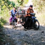 Warga dan seorang pedagang asongan tampak hati-hati mengendarai sepeda motornya karena kondisi jalan yang rusak parah.