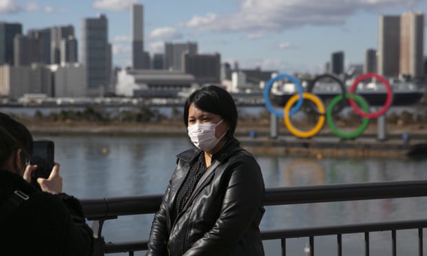 Seorang turis yang mengenakan topeng berpose untuk foto dengan cincin Olimpiade di latar belakang, di distrik Odaiba Tokyo. foto: theguardian